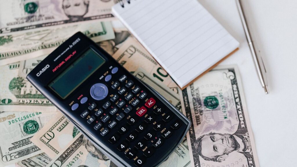 A calculator lying on cash, with a notepad and a pen.