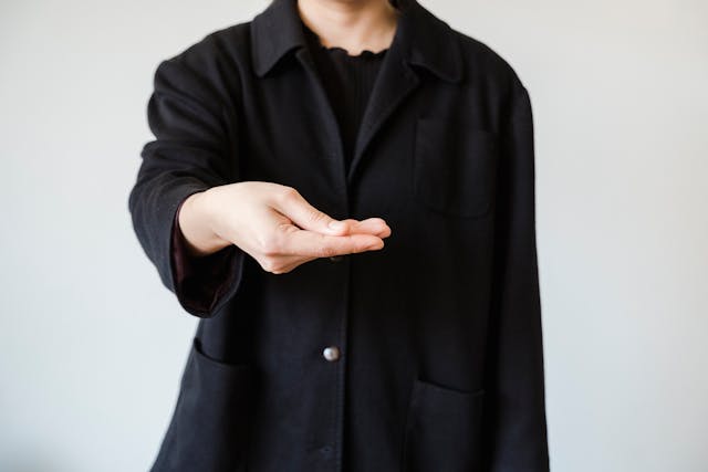 A man in a black outfit with his hand outstretched to the viewer as if expecting to receive something.  His head is cropped.